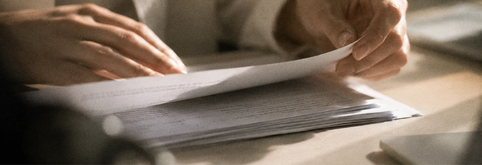 Hands leafing through a stack of printed documents on a desk in warm side light