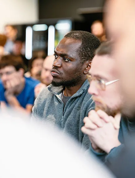 Teilnehmer eines Team-Events sitzt im Publikum und hört aufmerksam zu, während weitere Personen im Hintergrund zu sehen sind.