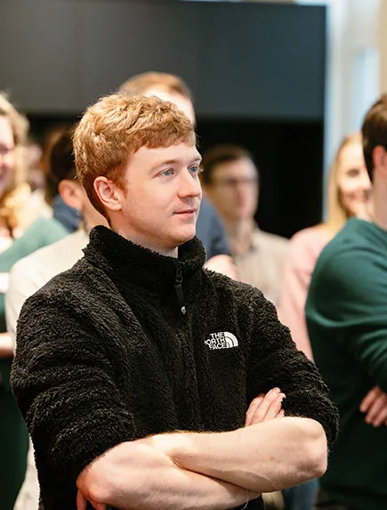 Teilnehmer steht mit verschränkten Armen im Publikum und hört einer Präsentation zu.